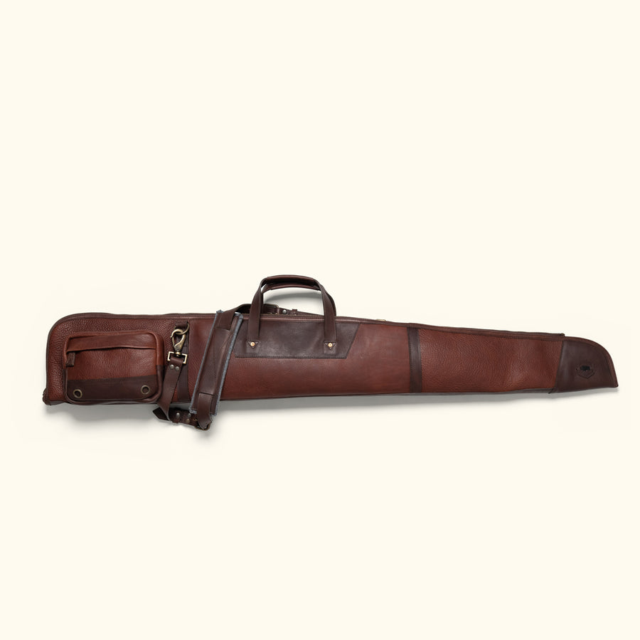 Rustic leather shotgun case draped over a barbed wire fence, with a backdrop of open fields.